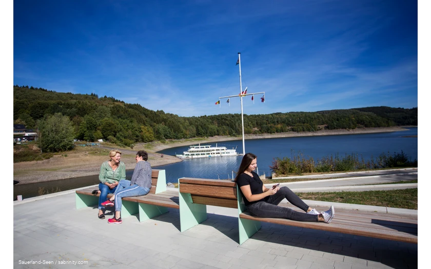 In the foreground, three women are sitting on benches, two are talking and one is on her cell phone. In the background is Lake Bigee with the boat.