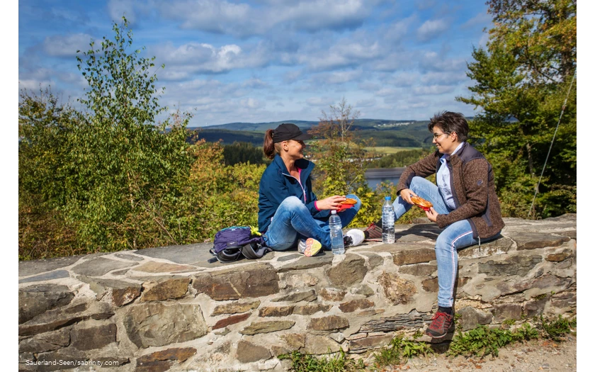 Zwei Frauen machen Rast auf einer Mauer mit Seeblick