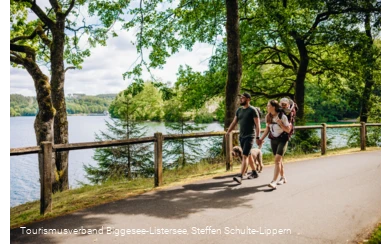 Ein Paar mit Hund und Kind in einer Trage wandert den Bigge-/Listersee entlang. Im Hintergrund ist die Staumauer zu erkennen.