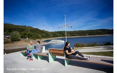 In the foreground, three women are sitting on benches, two are talking and one is on her cell phone. In the background is Lake Bigee with the boat.