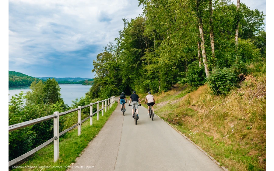 Drei Männer fahren entlang des Bigge-/Listersees auf ihren Fahrrädern weg von der Kamera. Der Himmel im Hintegrund ist wolkenverhangen.