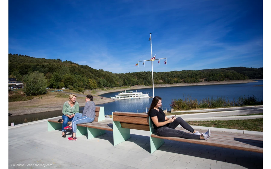 In the foreground, three women are sitting on benches, two are talking and one is on her cell phone. In the background is Lake Bigee with the boat.