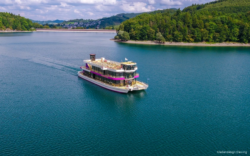 Die EMS Hennesee schippert über den Hennesee. Im Hintergrund ist die Staumauer und Teile von Meschede zu sehen.
