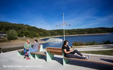 Im Vordergrund sitzen drei Frauen auf Bänken, zwei unterhalten sich und eine ist am Handy. Im Hintergrund ist der Bigeesee mit dem Schiff.