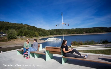 Im Vordergrund sitzen drei Frauen auf Bänken, zwei unterhalten sich und eine ist am Handy. Im Hintergrund ist der Bigeesee mit dem Schiff.