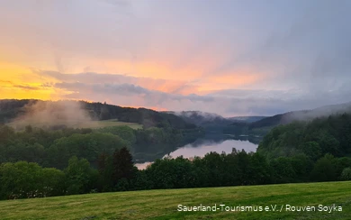 Über dem Hennesee und den Bäumen steigt Nebel auf und der Himmel kreiert mit Wolken und der unterhehenden Sonne ein Farbspektakel.