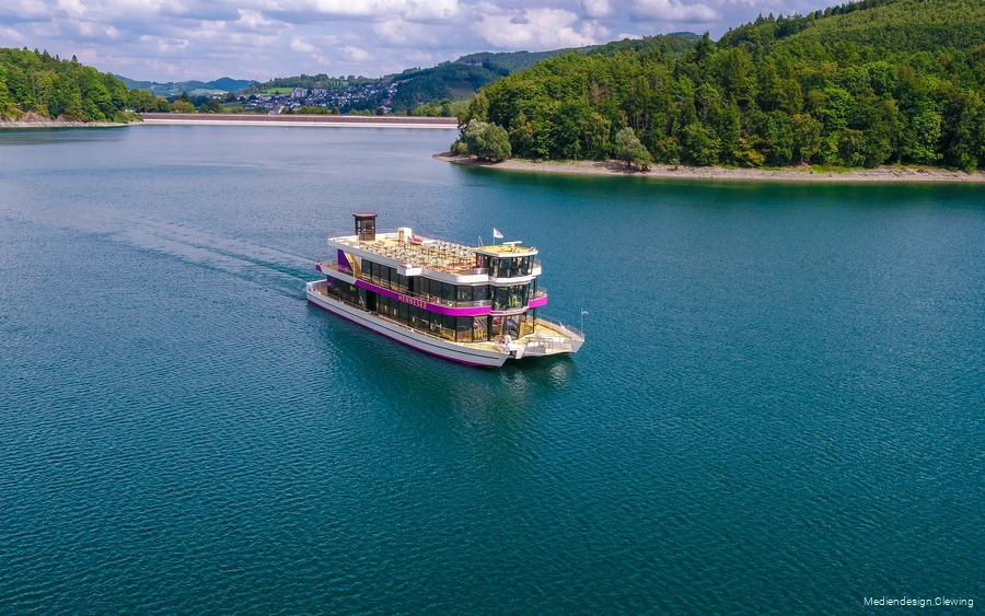 Die EMS Hennesee schippert über den Hennesee. Im Hintergrund ist die Staumauer und Teile von Meschede zu sehen.