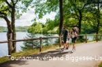 Ein Paar mit Hund und Kind in einer Trage wandert den Bigge-/Listersee entlang. Im Hintergrund ist die Staumauer zu erkennen.