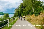 Drei Männer fahren entlang des Bigge-/Listersees auf ihren Fahrrädern weg von der Kamera. Der Himmel im Hintegrund ist wolkenverhangen.