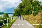 Drei Männer fahren entlang des Bigge-/Listersees auf ihren Fahrrädern weg von der Kamera. Der Himmel im Hintegrund ist wolkenverhangen.