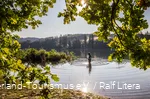 Ein Angler steht mit seiner Route im See. Die Sonne glitzert auf dem Wasser und die Blätter einer Eiche leuchten grün.